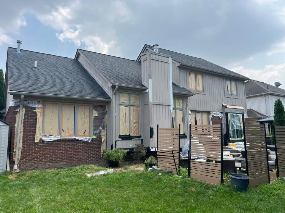 The same two-story home before painting: worn taupe board-and-batten siding with windows and doors papered off and masked for spraying